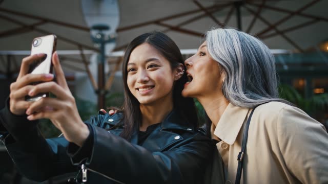 Asian-middle-aged-mother-and-adult-daughter-taking-selfie-by-smartphone-and-smiling-while-posing-near-terrace-of-cafe