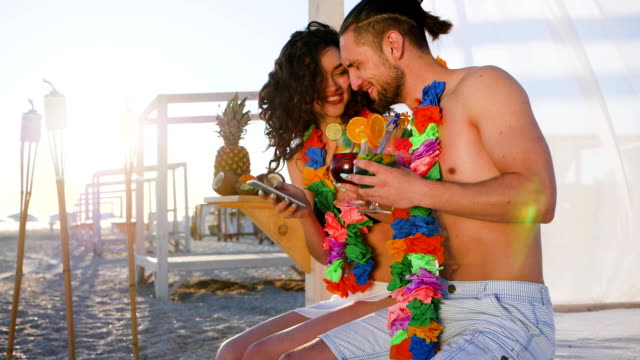 comunicación-móvil,-niña-sentado-con-chico-en-la-playa-y-sosteniendo-en-las-manos-celular,-hermosa-pareja,-jóvenes-de-lei-de-Hawaii-en-cuello-bebida-coctel-ver-las-redes-sociales