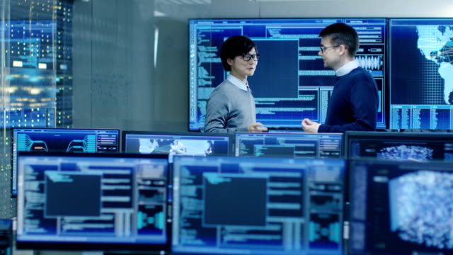 In-the-System-Control-Room-Project-Manage-and-IT-Engineer-Have-Discussion,-they're-surrounded-by-Multiple-Monitors-with-Graphics.-They-Work-in-a-Data-Center-on-Data-Mining,-AI-and-Neural-Networking.