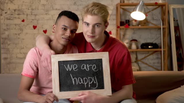 A-sad-international-gay-couple-is-sitting-on-the-couch-and-holding-a-sign.-We-are-not-happy.-Look-at-the-camera.-Home-comfort-on-the-background.-60-fps