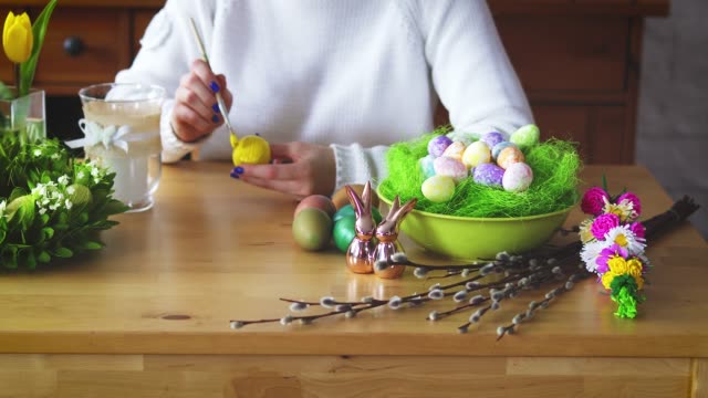 woman-paints-a-yellow-egg-on-the-table-with-Easter-decorations