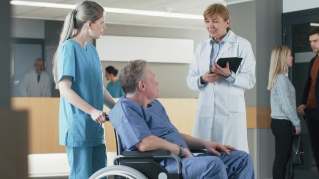 In-der-Klinik-Lobby,-Krankenschwester-schiebt-älteren-Patienten-im-Rollstuhl,-zwar-Arzt-spricht-mit-ihnen-mittels-Tablet-Computer.-Sauberes,-neues-Krankenhaus-mit-medizinischen-Fachpersonal.-Slow-Motion.