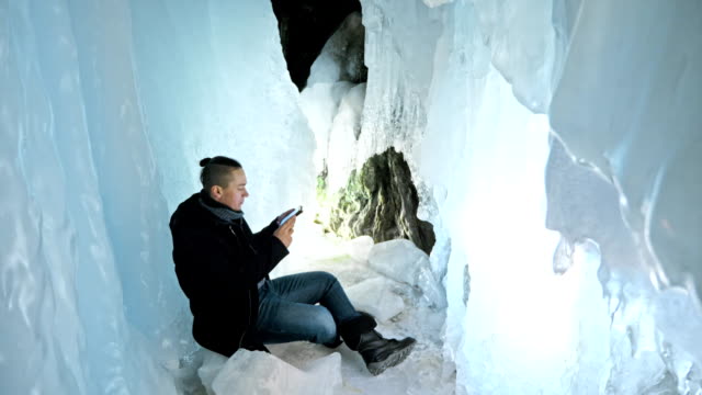Man-is-sit-on-the-internet-in-tablet-in-an-ice-cave.-Around-the-mysterious-beautiful-ice-grotto.-User-communicates-in-social-networks,-takes-photo-and-selfie-on-the-tablet.-He-crazy-strange-hipster.