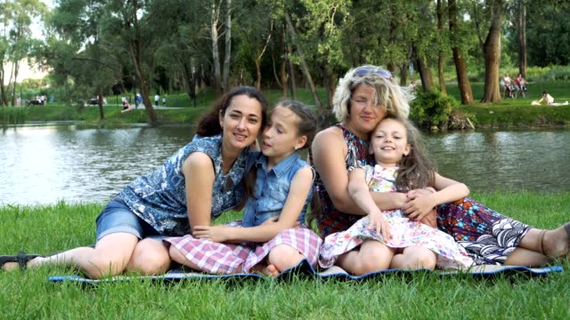 Happy-female-family-playing-in-the-Park.-Two-mothers-and-two-daughters-sit-on-the-lawn-by-the-river-at-sunset-laughing,-hugging-and-kissing.