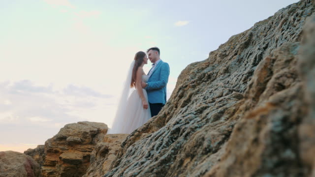 Beautiful-young-wedding-couple-standing-on-sea-shore-with-rocks.-Newlyweds-spend-time-together:-embrace,-kiss-and-care-for-each-other.-Camera-moves-from-behind-rock