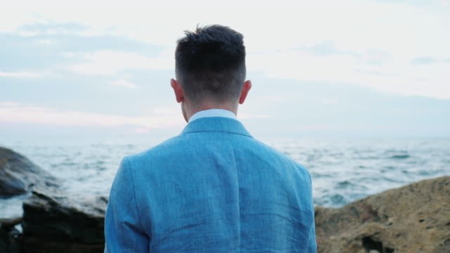 Portrait-of-young-attractive-business-man-or-groom-stands-on-sea-shore-with-rocks-and-watching-beautiful-sunset-or-sunrise-alone.-4k.-Slow-motion