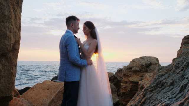 Beautiful-young-wedding-couple-standing-on-sea-shore-with-rocks.-Newlyweds-spend-time-together:-embrace,-kiss-and-care-for-each-other.-Love-concept.-Bride-smiling-to-groom