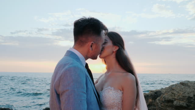 Beautiful-young-wedding-couple-standing-on-sea-shore-with-rocks.-Newlyweds-spend-time-together:-embrace,-kiss-and-care-for-each-other.-Love-concept