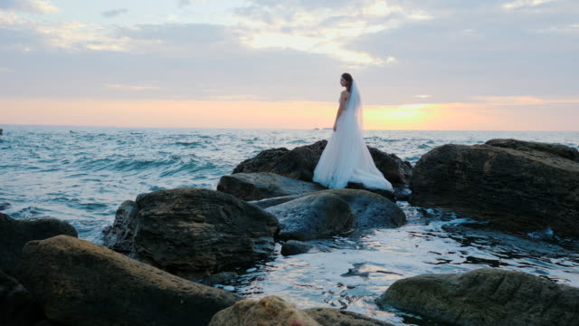 Girl-in-wedding-luxury-dress-posing-on-sea-shore.-Bride-on-a-rocks.-Beautiful-waves-near-to-her.-Woman-enjoying-happy-moments-with-nature.-Sunrise-or-sunset-time.-Slow-motion