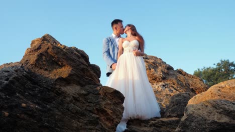 Schöne-junge-Hochzeitspaar-steht-am-Ufer-des-Meeres-mit-Felsen.-Brautpaar-gemeinsam-Zeit-verbringen:-umarmen,-küssen-und-füreinander.-Kamerafahrten-hinter-Felsen