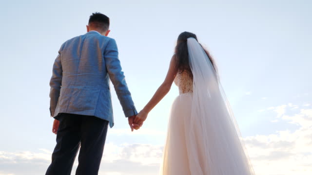 Unrecognizable-wedding-couple-holding-hands-on-sunset-or-sunrise-background.-Bride-and-groom-standing-outdoor-near-sea.-Sun-flares.-Slow-motion