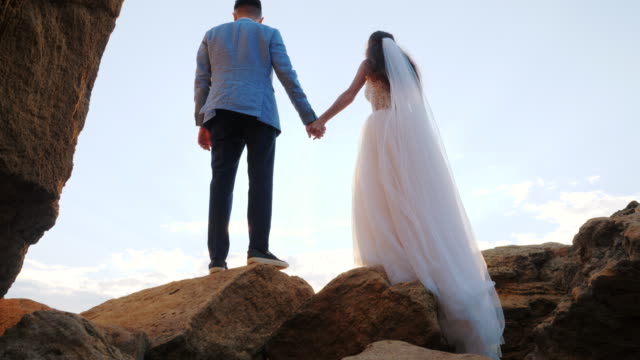 Unrecognizable-wedding-couple-holding-hands-on-sunset-or-sunrise-background.-Bride-and-groom-standing-outdoor-near-sea.-Sun-flares.-Slow-motion