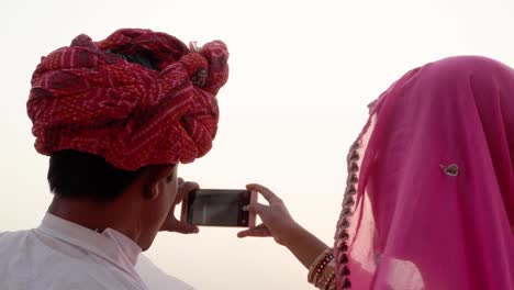 Indian-couple-looking-back-at-the-camera-and-taking-photos-of-sunset-at-holy-Pushkar-lake,-Rajasthan