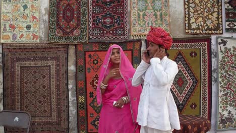 Indian-couple-in-traditional-clothes-enter-the-frame-and-attend-a-phone-call-with-traditional-carpets-hanging-in-the-backdrop