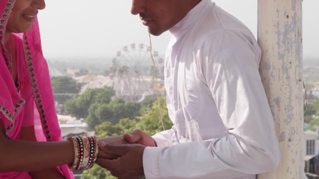 Romantic-couple-dating-at-a-secret-location-with-view-of-a-carnival-in-the-backdrop-in-pink-sari,-red-turban-and-white-kurta