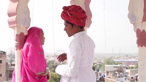 Tilt-down-to-dating-girl-and-boy-under-a-rooftop-canopy-chattri-talking-in-Rajasthan,-India-in-pink-sari-and-red-turban-and-white-kurta