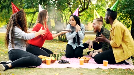 Friends-are-congratulating-Asian-woman-on-birthday-giving-cake-making-surprise,-girl-is-blowing-candles,-smiling-and-clapping-hands-during-outdoor-party-in-park.