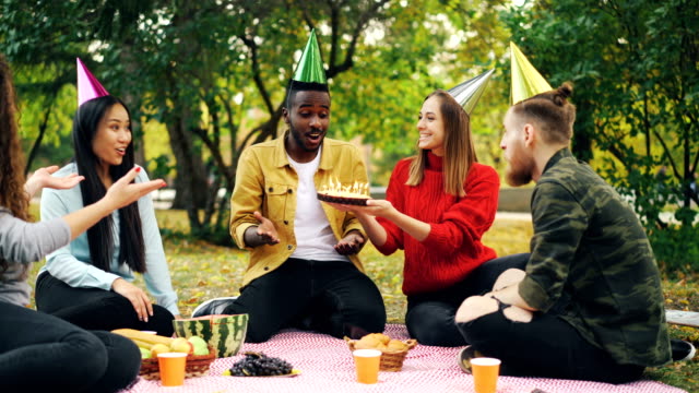Afroamerikanische-bärtigen-Kerl-ist-im-Park-bläst-Kerzen-auf-Kuchen-und-lachen-genießen-Überraschung-Geburtstagsfeier-mit,-seine-Freunde-sind-Händeklatschen.