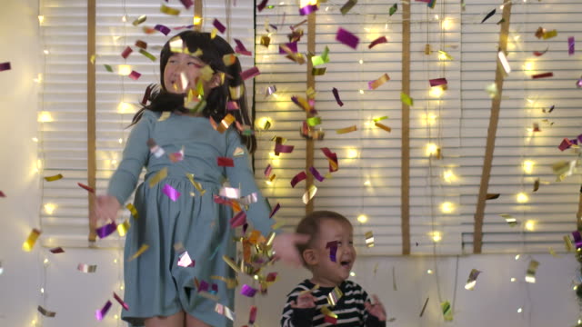 Two-happy-asian-children-plays-with-confetti-and-have-fun-jumping-in-living-room-in-slow-motion.