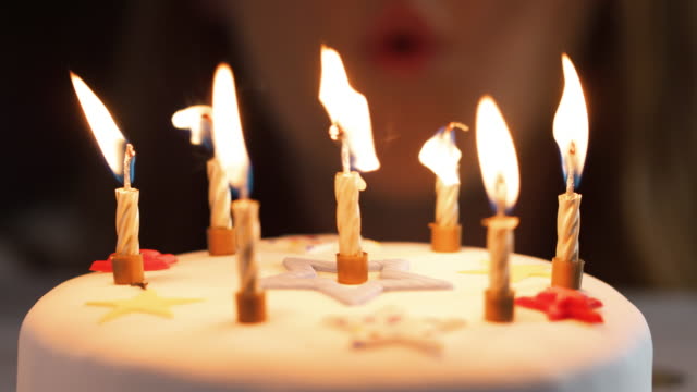 Close-up-of-a-woman-blowing-out-seven-lit-candles-on-a-white,-decorated-birthday-cake,-a-party-blower-beside-it,-bokeh-lights-in-the-background,-detail