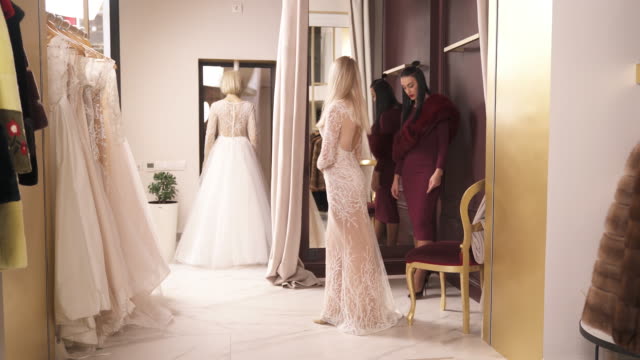 A-young-blonde-in-a-long-veiling-fancy-dress-is-turning-around-in-front-of-the-mirror-and-talking-to-the-designer-standing-to-the-left.-They-are-in-a-wedding-dress-atelier.