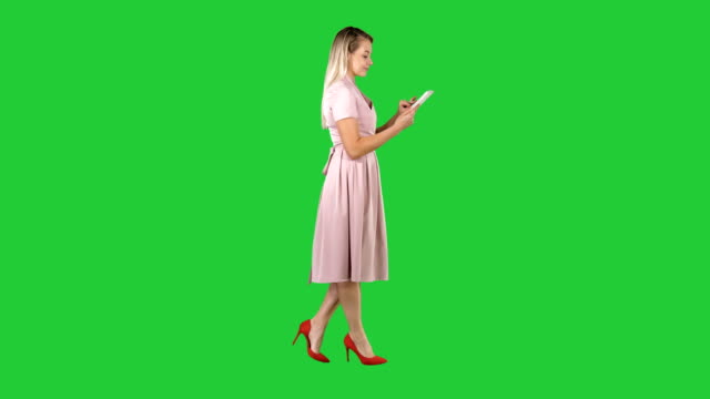 Blonde-in-pink-dress-walking-and-using-tablet-on-a-Green-Screen,-Chroma-Key