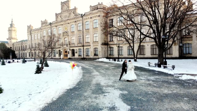 winter-wedding.-Aerial-view-of-newlywed-couple-in-wedding-dresses-are-dancing-wedding-dance-in-a-snow-covered-park,-against-the-background-of-ancient-architecture-and-paving-stones