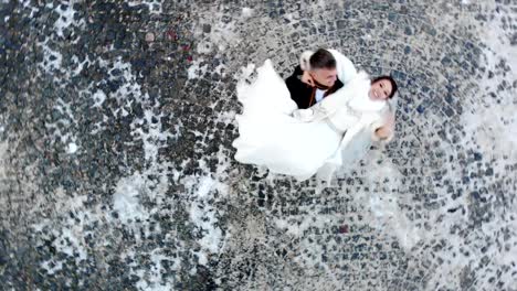 winter-wedding.-Aerial-view-of-newlywed-couple-in-wedding-dresses-are-dancing-wedding-dance-in-a-snow-covered-park,-against-the-background-of-paving-stones