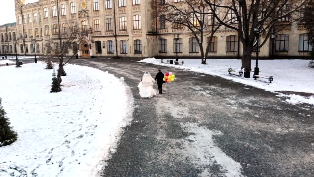 winter-wedding.-Aerial-view,-newlywed-couple-in-wedding-dresses-are-runing-with-colorful-balloons-through-the-snow-covered-park,-against-the-background-of-ancient-architecture-and-paving-stones
