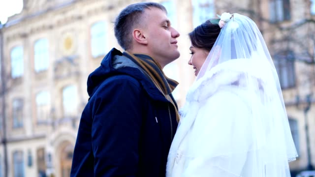 winter-wedding.-newlywed-couple-in-wedding-dresses.-groom-gently-kisses-the-bride-on-the-forehead,-hug.-they-are-happy,-smiling-at-each-other.-background-of-ancient-architecture,-snow-covered-park
