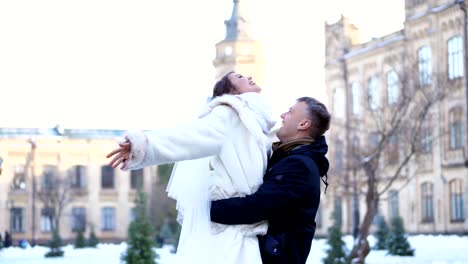 winter-wedding.-newlywed-couple-in-wedding-dresses.-groom-holds-bride-in-his-arms,-spinning.-they-are-happy,-smiling-at-each-other.-background-of-ancient-architecture,-snow-covered-park