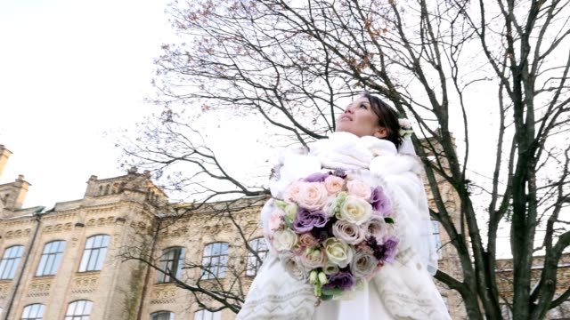 portrait-of-a-beautiful-bride,-in-a-white-dress,-veil-and-fur-coat.-holding-a-wedding-bouquet.-against-the-background-of-ancient-architecture,-the-sky,-trees-without-leaves.-winter-wedding
