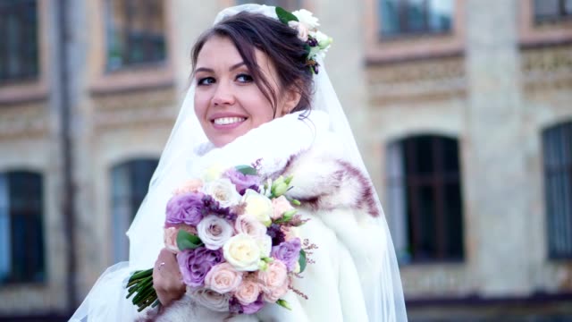 portrait-of-a-beautiful-bride,-in-a-white-dress,-veil-and-fur-coat.-bride-is-smiling-happily,-holding-a-wedding-bouquet.-against-the-background-of-ancient-architecture,-winter-wedding