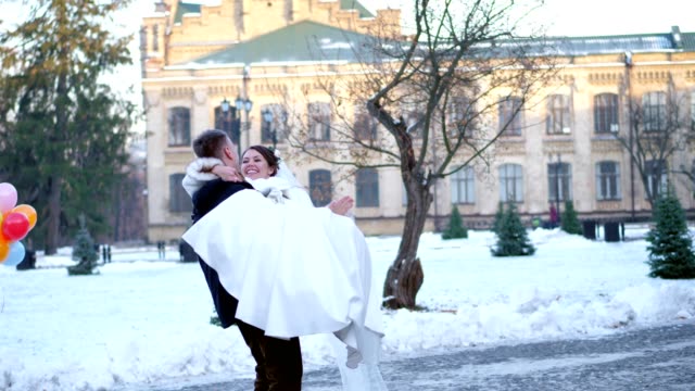 winter-wedding.-newlywed-couple-in-wedding-dresses.-groom-holds-bride-in-his-arms,-spinning.-they-are-happy,-smiling-to-each-other.-background-of-ancient-architecture,-snow-covered-park