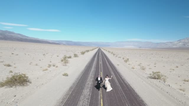 Bride-and-groom-running-on-empty-desert-road-at-Death-Valley,-USA