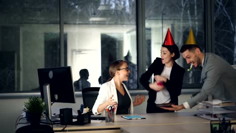 Pretty-blonde-is-working-in-modern-office-when-her-colleagues-are-bringing-birthday-cake,-present-and-party-hats,-girl-is-opening-gift-box-and-enjoying-congratulations