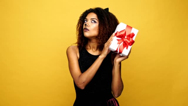 Young-happy-african-woman-cat-with-pretty-make-up-shaking-present-and-smiling-isolated-over-yellow