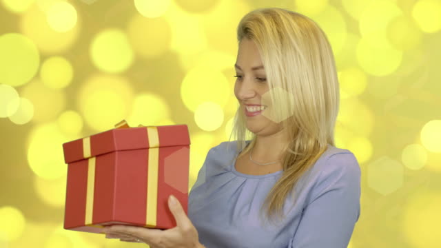 Excited-cheerful-attractive-young-woman-in-blue-dress-opening-gift-over-gold-background