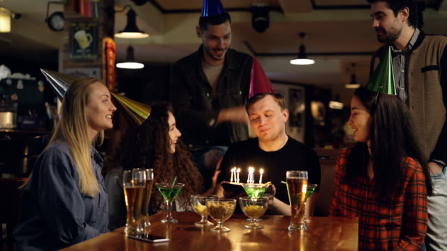 Best-friends-are-congratulating-young-man-on-birthday,-he-is-blowing-out-candles-on-cake-and-thanking-his-mates-for-great-party.-Funny-holiday-with-friends-concept.