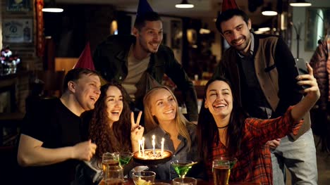 Happy-attractive-students-in-birthday-party-hats-are-recording-video-with-smartphone-sitting-at-table-in-bar.-They-are-posing-with-cake,-talking-and-laughing.