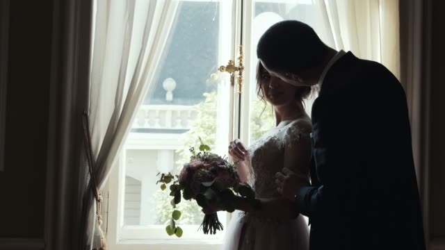 Marrying-couple-embrace-and-kiss-standing-near-huge-window-in-room