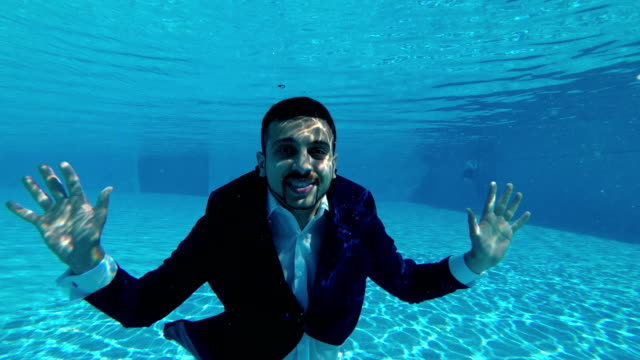 A-man-in-a-blue-suit-and-a-white-shirt-swims-and-poses-underwater-in-the-pool.-He-looks-at-the-camera,-waves-his-hands-and-smiles.