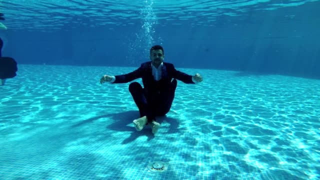 The-happy-groom-in-a-suit-and-a-white-shirt-goes-down-underwater-to-the-bottom-of-the-pool.-He-sits-at-the-bottom-in-a-Lotus-position,-looks-at-the-camera-and-smiles.-Slow-motion.-Action-camera-underwater.