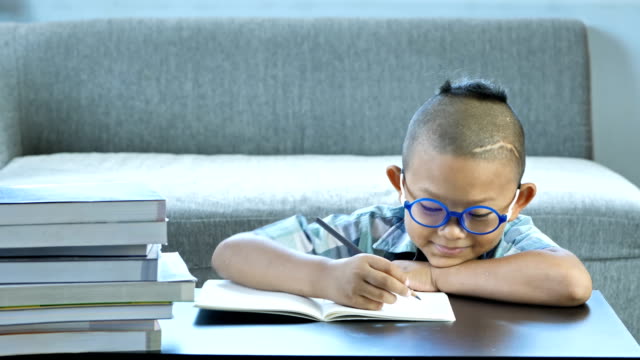 Boy-with-disability,-brain-disorders-And-Left-eye-is-not-visible-from-brain-surgery.-He-is-thinking-and-write-into-book,-Happy-smile-at-home.-education-concept