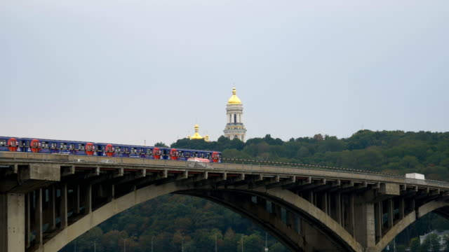 Train-travels-over-the-bridge-against-the-sky-and-the-domes-of-the-church.-City-subway-left-outside.-Public-transport-on-the-move.-Cars-go-over-the-bridge-over-the-river.