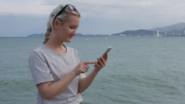 Schöne-schlanke-Frau-mit-einem-Zopf-Blondine-in-einem-grauen-T-shirt-stehend-und-mit-Smartphone-über-Hintergrund-Meer-und-Inseln.-Bildschirm-berühren-Mädchen