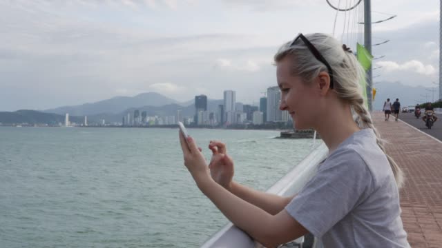 Schöne-schlanke-Frau-mit-einem-Zopf-Blondine-in-einem-grauen-T-shirt-auf-der-Brücke-stehend-und-mit-Smartphone-über-Hintergrund-Meer-und-Stadt.-Bildschirm-berühren-Mädchen