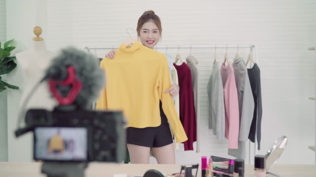 Asian-fashion-female-blogger-online-influencer-holding-shopping-bags-and-lots-of-clothes-on-clothes-rack-for-recording-new-fashion-video-broadcast-live-video-to-social-network-by-internet-at-home.