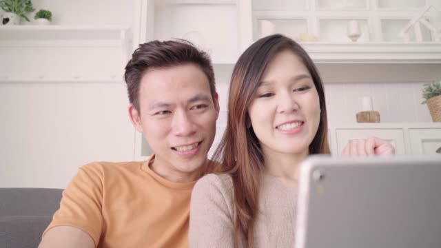 Asian-paar-mit-Tablet-Videoanruf-mit-Freund-im-Wohnzimmer-zu-Hause,-süßes-Paar-Liebe-Moment-beim-liegen-auf-dem-Sofa,-wenn-zu-Hause-entspannt-genießen.-Lifestyle-paar-entspannen-zu-Hause-Konzept.
