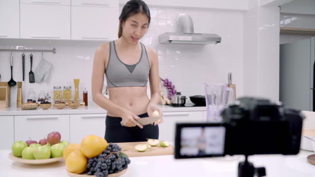 Blogger-deportiva-mujer-asiática-con-cámara-de-grabación-cómo-realizar-el-video-de-jugo-de-manzana-para-su-abonado,-mujer-frutas-orgánicas-haciendo-jugo-de-manzana-por-ella-misma-en-casa.-Concepto-de-comida-sana.